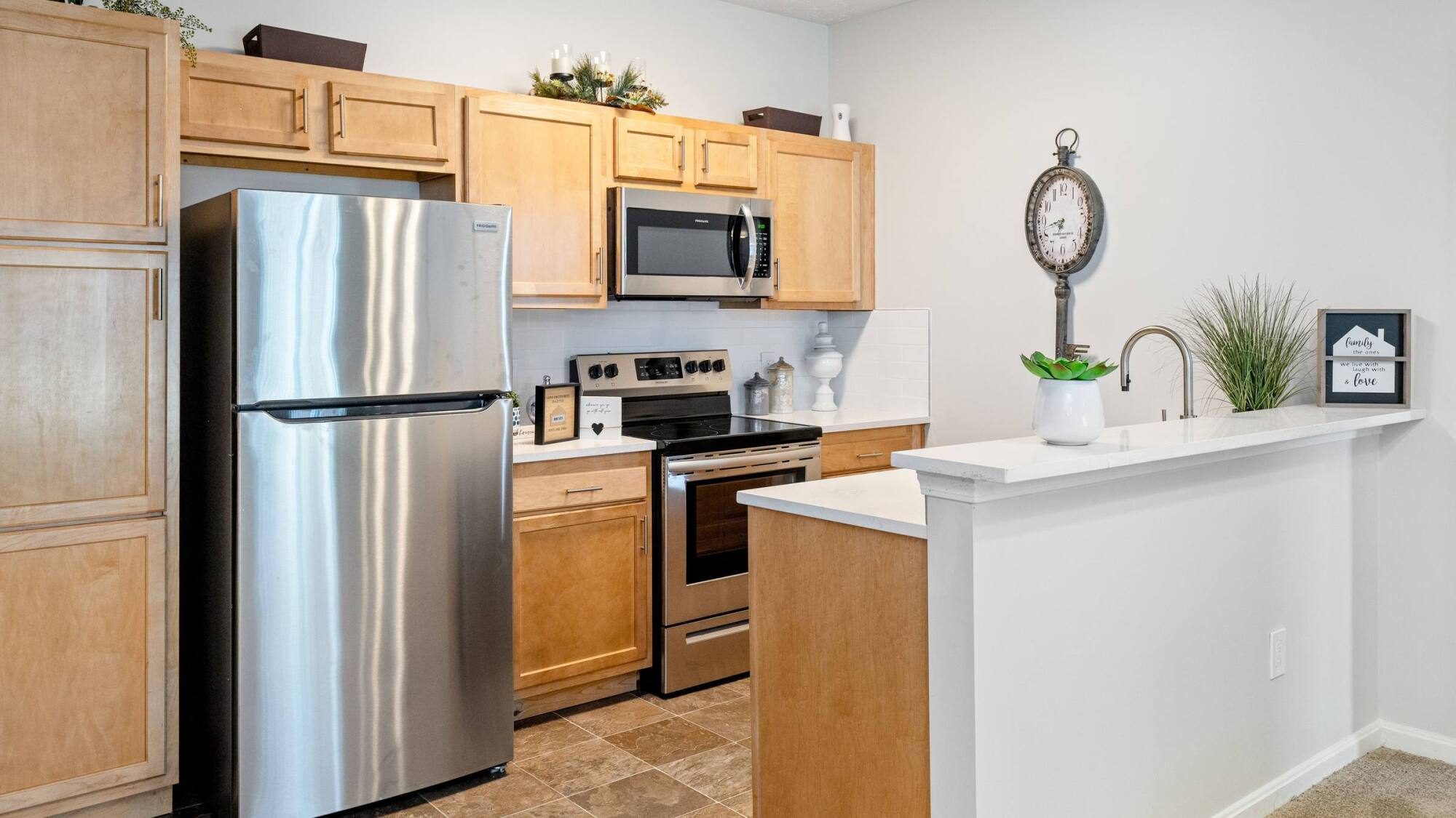 Modern kitchen with wood cabinets, stainless steel appliances, a wall clock, and white countertops with decorative plants.